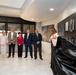 A formal unveiling ceremony is taking place in what appears to be a hall of fame or memorial corridor. A woman in a colorful dress is pulling back a black drape to reveal framed portraits and plaques mounted on a dark wall, while six attendees stand and observe. The wall displays what appears to be a 'Soldier's Creed' panel and multiple framed photographs with biographical plaques, suggesting a military or institutional recognition display.