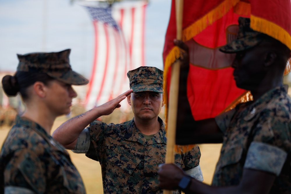 Three U.S. Marines in MARPAT digital camouflage uniforms are participating in a formal military ceremony. A central Marine is rendering a salute while a second Marine holds a red and gold unit guidon or colors. A large American flag is visible in the blurred background.