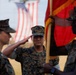 Three U.S. Marines in MARPAT digital camouflage uniforms are participating in a formal military ceremony. A central Marine is rendering a salute while a second Marine holds a red and gold unit guidon or colors. A large American flag is visible in the blurred background.