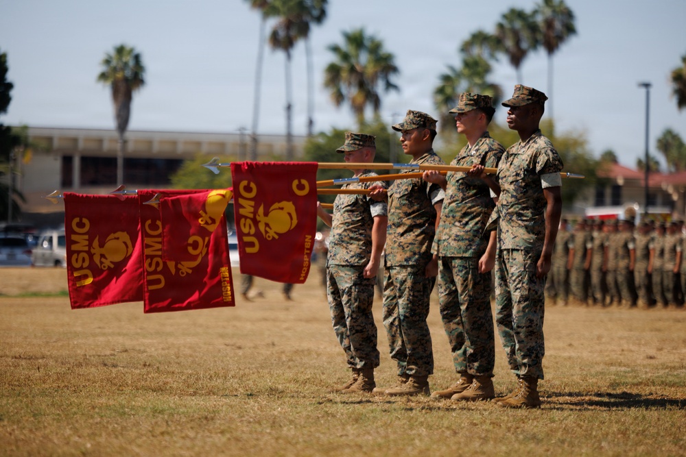 A formal United States Marine Corps (USMC) military ceremony is taking place on a parade ground, likely at a base in Southern California given the palm trees and architecture. Four Marines in woodland MARPAT camouflage uniforms are holding red USMC guidon flags with gold Eagle, Globe, and Anchor emblems. A formation of additional Marines stands at attention in the background.