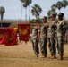 A formal United States Marine Corps (USMC) military ceremony is taking place on a parade ground, likely at a base in Southern California given the palm trees and architecture. Four Marines in woodland MARPAT camouflage uniforms are holding red USMC guidon flags with gold Eagle, Globe, and Anchor emblems. A formation of additional Marines stands at attention in the background.
