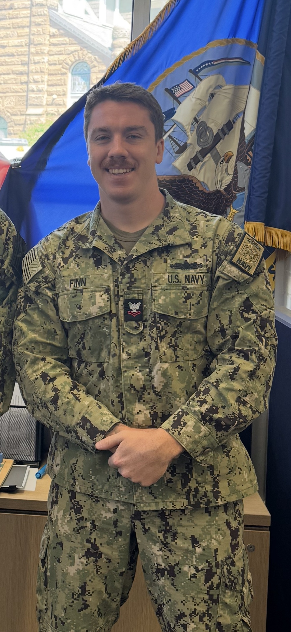 A U.S. Navy sailor in Navy Working Uniform (NWU Type III) digital camouflage stands smiling in what appears to be a recruiting or administrative office. The name tape reads 'FINN' and the branch tape reads 'U.S. NAVY,' with visible rank insignia indicating a Petty Officer Second Class. A U.S. Navy ceremonial flag with gold fringe is visible in the background.