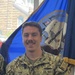 A U.S. Navy sailor in Navy Working Uniform (NWU Type III) digital camouflage stands smiling in what appears to be a recruiting or administrative office. The name tape reads 'FINN' and the branch tape reads 'U.S. NAVY,' with visible rank insignia indicating a Petty Officer Second Class. A U.S. Navy ceremonial flag with gold fringe is visible in the background.