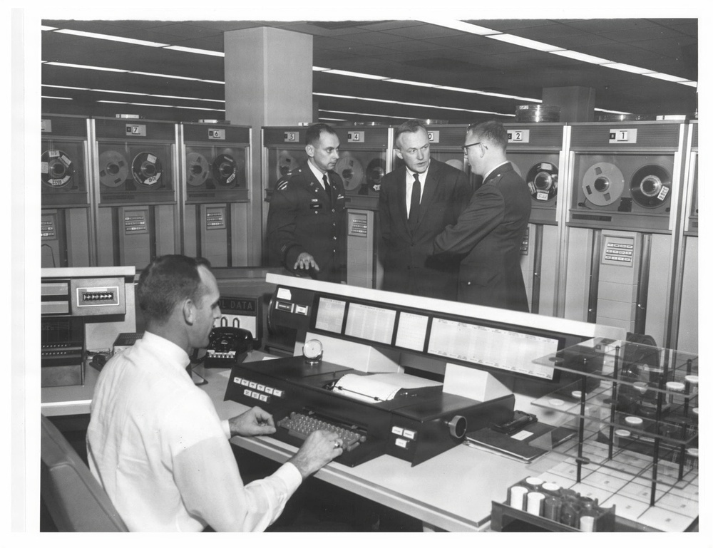 A black-and-white photograph depicting what appears to be a military or government computer operations center, likely from the 1960s. One operator is seated at a large console with a keyboard and telephone, while three men — two in military uniforms and one in a civilian suit — stand in conversation behind him. The background is lined with large reel-to-reel magnetic tape drive units numbered sequentially.