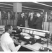 A black-and-white photograph depicting what appears to be a military or government computer operations center, likely from the 1960s. One operator is seated at a large console with a keyboard and telephone, while three men — two in military uniforms and one in a civilian suit — stand in conversation behind him. The background is lined with large reel-to-reel magnetic tape drive units numbered sequentially.