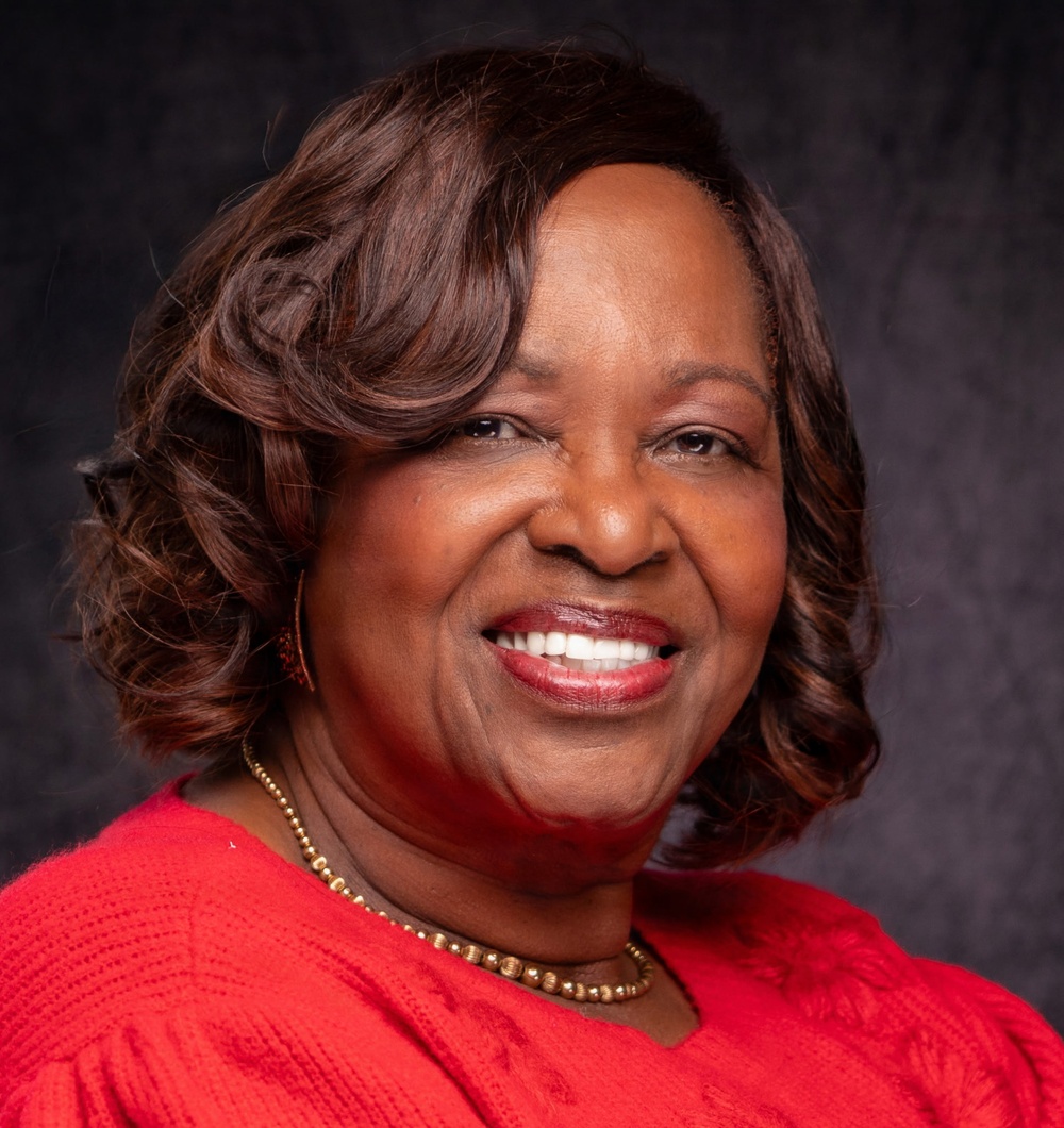A professional portrait photograph of a woman smiling warmly at the camera against a dark gray studio backdrop. She is dressed in a red top and wearing gold jewelry, suggesting this is a formal headshot or professional profile photo. The image is well-lit and high quality, consistent with professional photography.