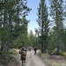 A group of military personnel in camouflage uniforms are conducting a patrol or road march along a dirt trail through a pine forest. The lead soldier is carrying what appears to be a large tube-launched weapon system (likely an AT4 or similar anti-tank rocket launcher) on their shoulder. The setting appears to be a training environment in a mountainous or high-elevation forested area.