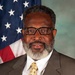 A formal portrait photograph of a middle-aged man wearing a brown pinstripe suit, white dress shirt, and gold patterned tie with a matching pocket square. He has short gray-streaked hair, a full gray beard, and black-framed glasses. An American flag is visible in the background to the left, suggesting this is an official or professional headshot.