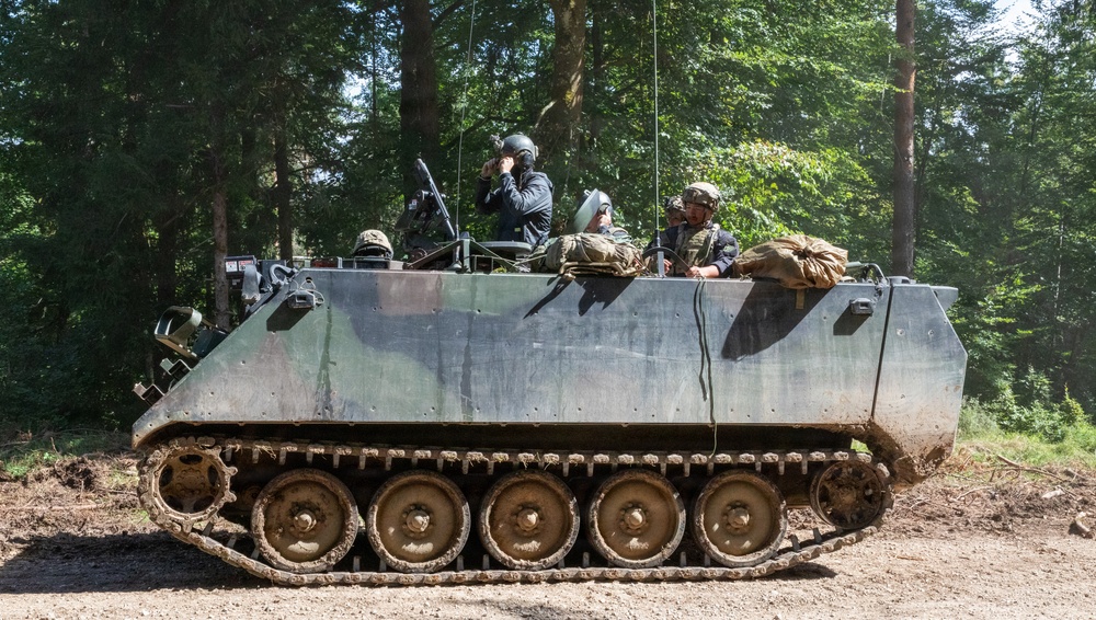 An armored personnel carrier (APC), identifiable as an M113 variant, is moving along a dirt track through a forested area. Several personnel are visible riding in the open top hatch, wearing military helmets and gear. The vehicle shows signs of active use with mud on the tracks and hull.