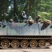 An armored personnel carrier (APC), identifiable as an M113 variant, is moving along a dirt track through a forested area. Several personnel are visible riding in the open top hatch, wearing military helmets and gear. The vehicle shows signs of active use with mud on the tracks and hull.