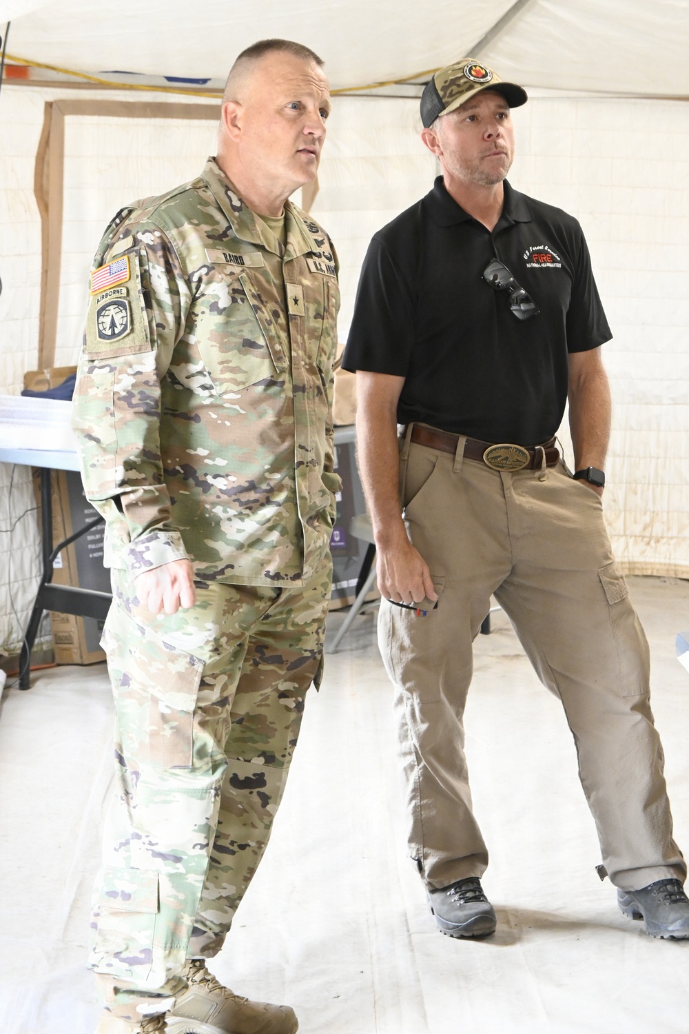 Two men stand inside what appears to be a tent or temporary structure. One man is wearing a U.S. Army OCP (Operational Camouflage Pattern) uniform with visible rank insignia (one star, indicating Brigadier General) and name tape reading 'BAIRD', along with Airborne and unit patches. The second man is wearing a black polo shirt with what appears to be a U.S. Forest Service Fire logo and a baseball cap, along with khaki cargo pants.
