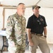 Two men stand inside what appears to be a tent or temporary structure. One man is wearing a U.S. Army OCP (Operational Camouflage Pattern) uniform with visible rank insignia (one star, indicating Brigadier General) and name tape reading 'BAIRD', along with Airborne and unit patches. The second man is wearing a black polo shirt with what appears to be a U.S. Forest Service Fire logo and a baseball cap, along with khaki cargo pants.