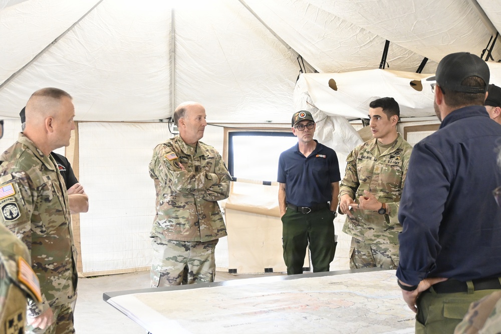 A group of U.S. Army personnel and at least one civilian are gathered inside a military command tent for what appears to be a briefing or planning session. They are standing around a table with maps spread out, suggesting operational or tactical planning activity. The setting is a standard military field tent with beige canvas walls and portable lighting.