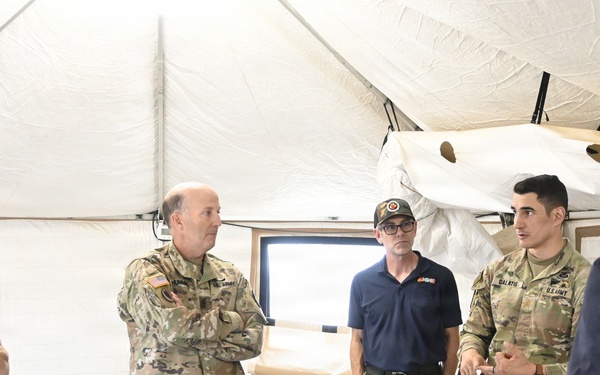A group of U.S. Army personnel and at least one civilian are gathered inside a military command tent for what appears to be a briefing or planning session. They are standing around a table with maps spread out, suggesting operational or tactical planning activity. The setting is a standard military field tent with beige canvas walls and portable lighting.