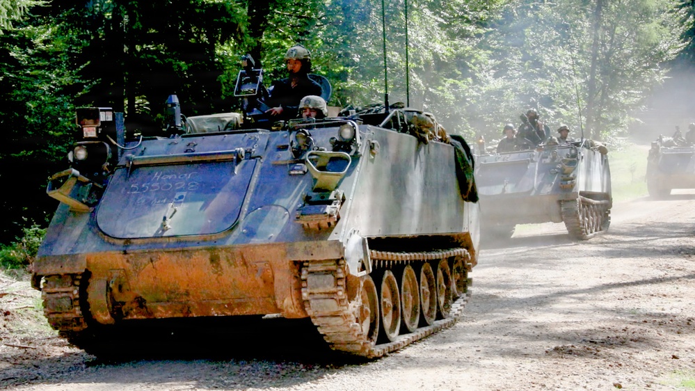 A column of M113 Armored Personnel Carriers (APCs) moves along a dirt forest road, likely during a military exercise or convoy operation. At least three vehicles are visible, with soldiers visible in the open hatches of each vehicle. The lead vehicle shows visible mud accumulation on its lower hull and tracks, suggesting recent off-road travel.