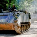 A column of M113 Armored Personnel Carriers (APCs) moves along a dirt forest road, likely during a military exercise or convoy operation. At least three vehicles are visible, with soldiers visible in the open hatches of each vehicle. The lead vehicle shows visible mud accumulation on its lower hull and tracks, suggesting recent off-road travel.