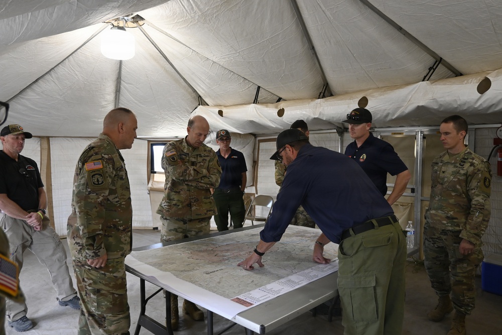 A group of military personnel and civilians are gathered inside a large military-style tent around a table with a large topographic or operational map spread out. One individual in civilian attire is leaning over the table pointing at specific locations on the map while others observe. The setting appears to be a forward operating base or emergency command post during a briefing or planning session.