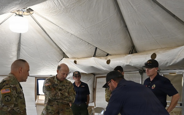 A group of military personnel and civilians are gathered inside a large military-style tent around a table with a large topographic or operational map spread out. One individual in civilian attire is leaning over the table pointing at specific locations on the map while others observe. The setting appears to be a forward operating base or emergency command post during a briefing or planning session.