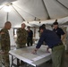 A group of military personnel and civilians are gathered inside a large military-style tent around a table with a large topographic or operational map spread out. One individual in civilian attire is leaning over the table pointing at specific locations on the map while others observe. The setting appears to be a forward operating base or emergency command post during a briefing or planning session.