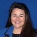 A professional headshot of a woman wearing a blue blazer over a black top, smiling against a blue background. She is wearing a lanyard with a 'TACOM' badge identifying her as 'Michele M. Moore' with the nickname 'M3'. A star-shaped pin or brooch is visible on her lapel.