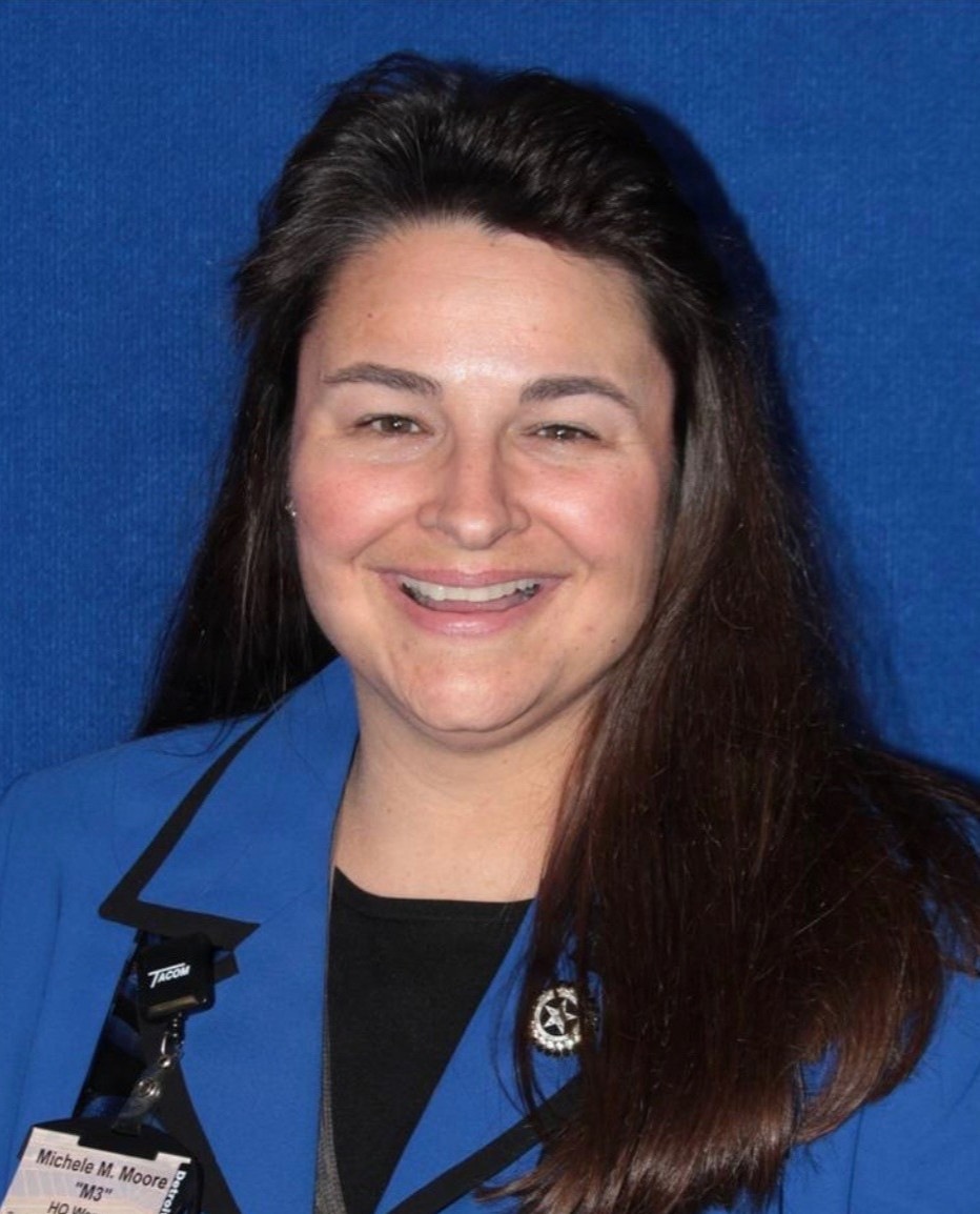 A professional headshot of a woman wearing a blue blazer over a black top, smiling against a blue background. She is wearing a lanyard with a 'TACOM' badge identifying her as 'Michele M. Moore' with the nickname 'M3'. A star-shaped pin or brooch is visible on her lapel.