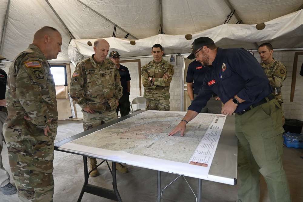 A group of U.S. Army soldiers in multicam uniforms and civilian emergency personnel are gathered inside a military tent around a large topographic map laid on a folding table. A civilian in dark blue uniform and green pants is pointing to specific locations on the map, appearing to brief the military personnel. The setting suggests a joint military-civilian emergency operations or incident command briefing.