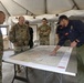 A group of U.S. Army soldiers in multicam uniforms and civilian emergency personnel are gathered inside a military tent around a large topographic map laid on a folding table. A civilian in dark blue uniform and green pants is pointing to specific locations on the map, appearing to brief the military personnel. The setting suggests a joint military-civilian emergency operations or incident command briefing.