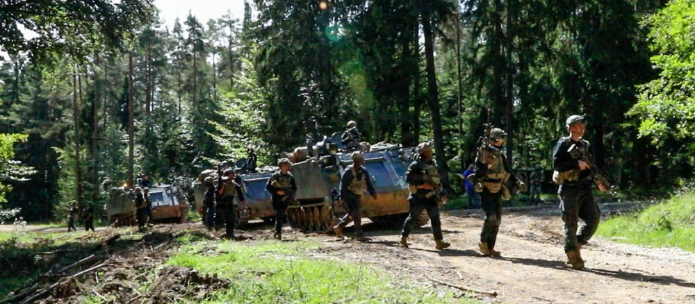 A column of military personnel and armored tracked vehicles moving along a dirt forest road. Soldiers in combat gear and helmets are advancing on foot alongside what appear to be M113 armored personnel carriers. The setting is a dense mixed forest under bright daylight conditions.