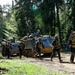 A column of military personnel and armored tracked vehicles moving along a dirt forest road. Soldiers in combat gear and helmets are advancing on foot alongside what appear to be M113 armored personnel carriers. The setting is a dense mixed forest under bright daylight conditions.