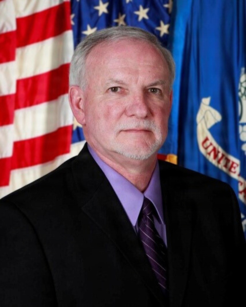 A formal portrait of an older male with gray hair and a goatee, wearing a black suit jacket, lavender dress shirt, and striped purple tie. He is posed in front of an American flag and what appears to be a blue organizational or agency flag with partial text reading 'UNITE'. The image appears to be an official government or agency headshot.
