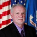 A formal portrait of an older male with gray hair and a goatee, wearing a black suit jacket, lavender dress shirt, and striped purple tie. He is posed in front of an American flag and what appears to be a blue organizational or agency flag with partial text reading 'UNITE'. The image appears to be an official government or agency headshot.