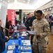A U.S. Army soldier named Wiggins is attending a military job fair or career expo, reviewing printed materials at a recruiter's table. A female representative in a blue blazer is speaking with him across a blue-draped table. The background shows a busy convention hall with multiple employer booths, including visible signage for Kiewit and Chick-fil-A.
