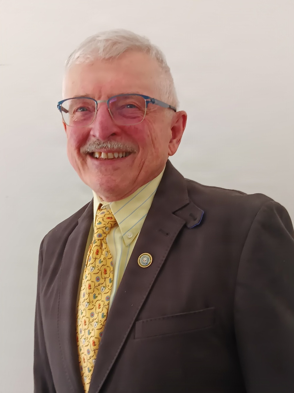 A formal portrait photograph of an older gentleman smiling warmly at the camera. He is dressed in professional attire including a dark brown blazer, yellow patterned tie, and light yellow striped dress shirt. A circular lapel pin is visible on his jacket.