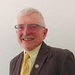 A formal portrait photograph of an older gentleman smiling warmly at the camera. He is dressed in professional attire including a dark brown blazer, yellow patterned tie, and light yellow striped dress shirt. A circular lapel pin is visible on his jacket.