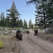 Three military personnel in full combat gear and camouflage uniforms are kneeling in tactical positions along a dirt trail in a forested, high-elevation terrain. They appear to be conducting a patrol halt or tactical movement exercise, with weapons raised or at the ready. The environment features pine trees, sagebrush, granite boulders, and a clear sky consistent with a mountainous western U.S. landscape.