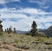 A wide open mountain landscape featuring a grassy, sagebrush-covered meadow with scattered conifer trees and distant mountain peaks under a partly cloudy blue sky. Two human figures are visible in the mid-ground, appearing small against the expansive terrain. The scene suggests a high-elevation wilderness area, likely in the western United States.
