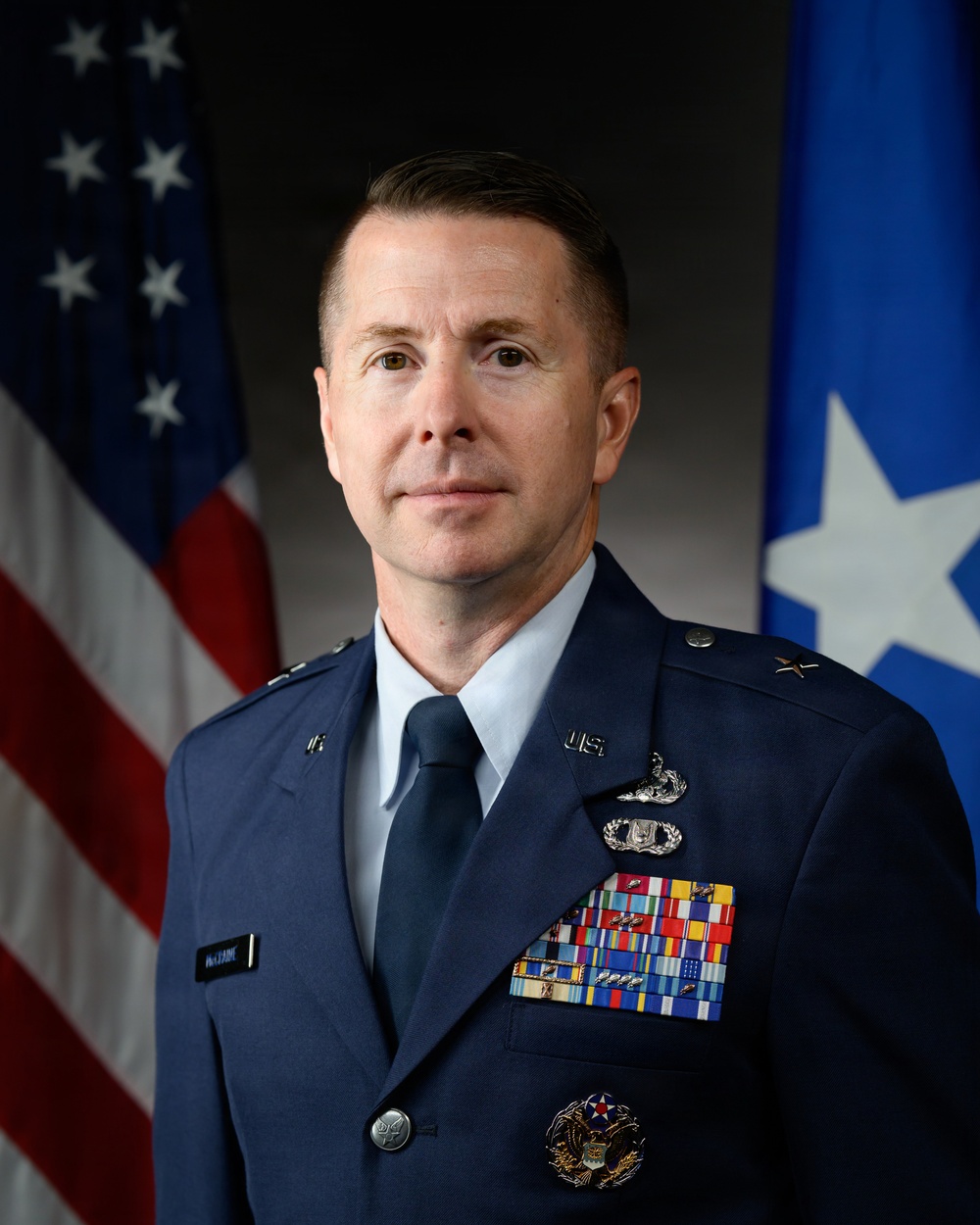 A formal military portrait of a U.S. Air Force officer in dress blue uniform. The subject displays a single star on each shoulder, indicating the rank of Brigadier General. The portrait is set against a backdrop featuring the U.S. flag and a blue general officer flag with a single white star.