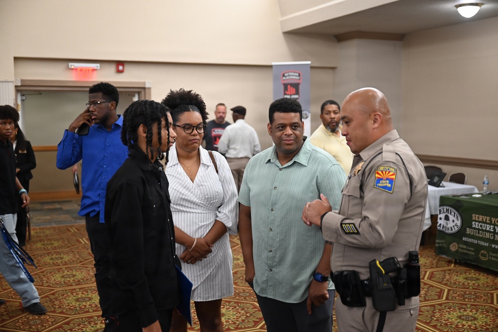 The image shows what appears to be a job fair or community outreach event held in a banquet or conference hall. An Arizona State Trooper in uniform is engaged in conversation with a small group of civilians, including adults and a younger individual. Various recruitment or informational banners are visible in the background.