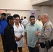 The image shows what appears to be a job fair or community outreach event held in a banquet or conference hall. An Arizona State Trooper in uniform is engaged in conversation with a small group of civilians, including adults and a younger individual. Various recruitment or informational banners are visible in the background.