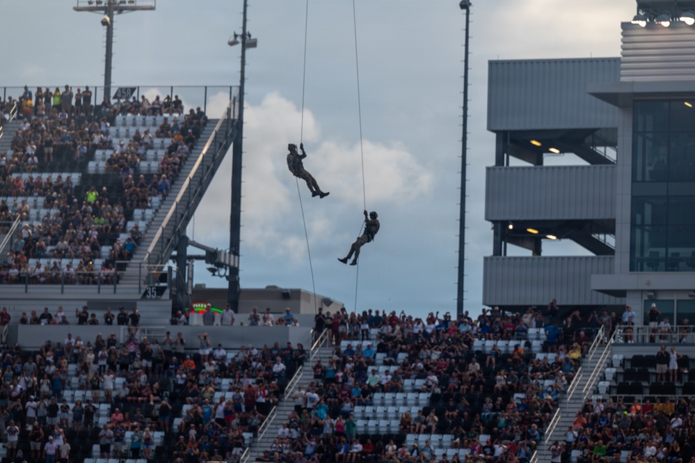 Two individuals in military-style gear are rappelling or fast-roping from ropes suspended high above a large stadium filled with spectators. The demonstration appears to be a pre-event or halftime military exhibition performed for a large crowd. The stadium structure, lighting towers, and multi-level press/luxury boxes are visible in the background.