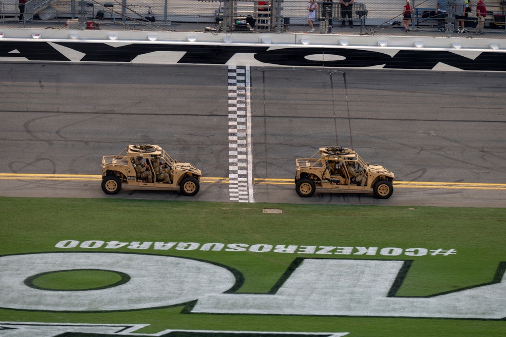 Two military tactical vehicles, appearing to be Oshkosh L-ATVs (Light Combat Tactical All-Terrain Vehicles) in tan/desert camouflage, are driving along the start/finish line at Daytona International Speedway. The vehicles are part of what appears to be a pre-race military display or ceremonial event. The infield grass displays the hashtag '#COKEZEROSUGAR400', identifying this as the Coke Zero Sugar 400 NASCAR race event.