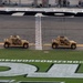 Two military tactical vehicles, appearing to be Oshkosh L-ATVs (Light Combat Tactical All-Terrain Vehicles) in tan/desert camouflage, are driving along the start/finish line at Daytona International Speedway. The vehicles are part of what appears to be a pre-race military display or ceremonial event. The infield grass displays the hashtag '#COKEZEROSUGAR400', identifying this as the Coke Zero Sugar 400 NASCAR race event.