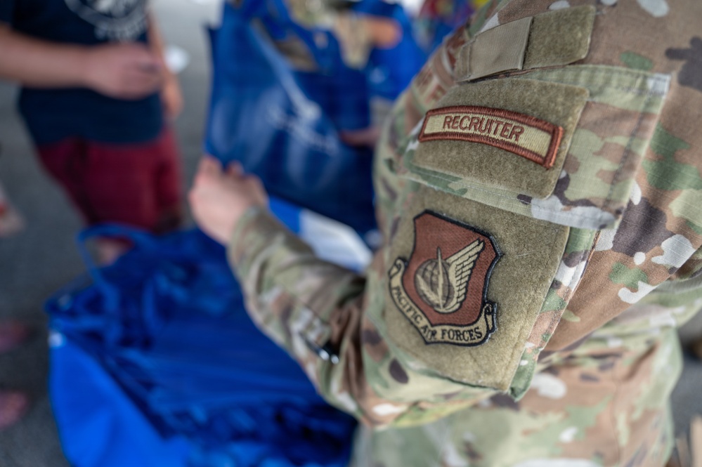 A U.S. Air Force recruiter in OCP (Operational Camouflage Pattern) uniform is handling blue bags or materials, likely at a recruiting or community outreach event. The soldier's sleeve clearly displays a 'RECRUITER' tab and a 'Pacific Air Forces' command patch. The scene suggests an interaction with civilians in an informal setting.
