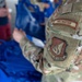 A U.S. Air Force recruiter in OCP (Operational Camouflage Pattern) uniform is handling blue bags or materials, likely at a recruiting or community outreach event. The soldier's sleeve clearly displays a 'RECRUITER' tab and a 'Pacific Air Forces' command patch. The scene suggests an interaction with civilians in an informal setting.