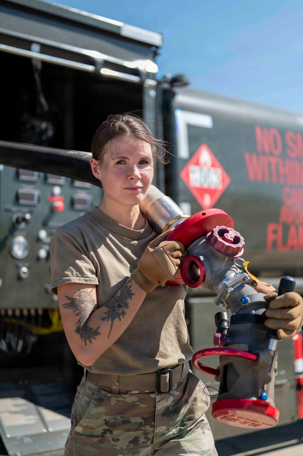 A female military service member in OCP (Operational Camouflage Pattern) uniform holds a large fuel nozzle/coupling assembly over her shoulder while standing in front of a military fuel tanker truck. She is wearing tan work gloves and has visible tattoos on her right forearm. The setting appears to be an outdoor military airfield or fuel operations area on a clear, sunny day.