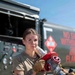 A female military service member in OCP (Operational Camouflage Pattern) uniform holds a large fuel nozzle/coupling assembly over her shoulder while standing in front of a military fuel tanker truck. She is wearing tan work gloves and has visible tattoos on her right forearm. The setting appears to be an outdoor military airfield or fuel operations area on a clear, sunny day.