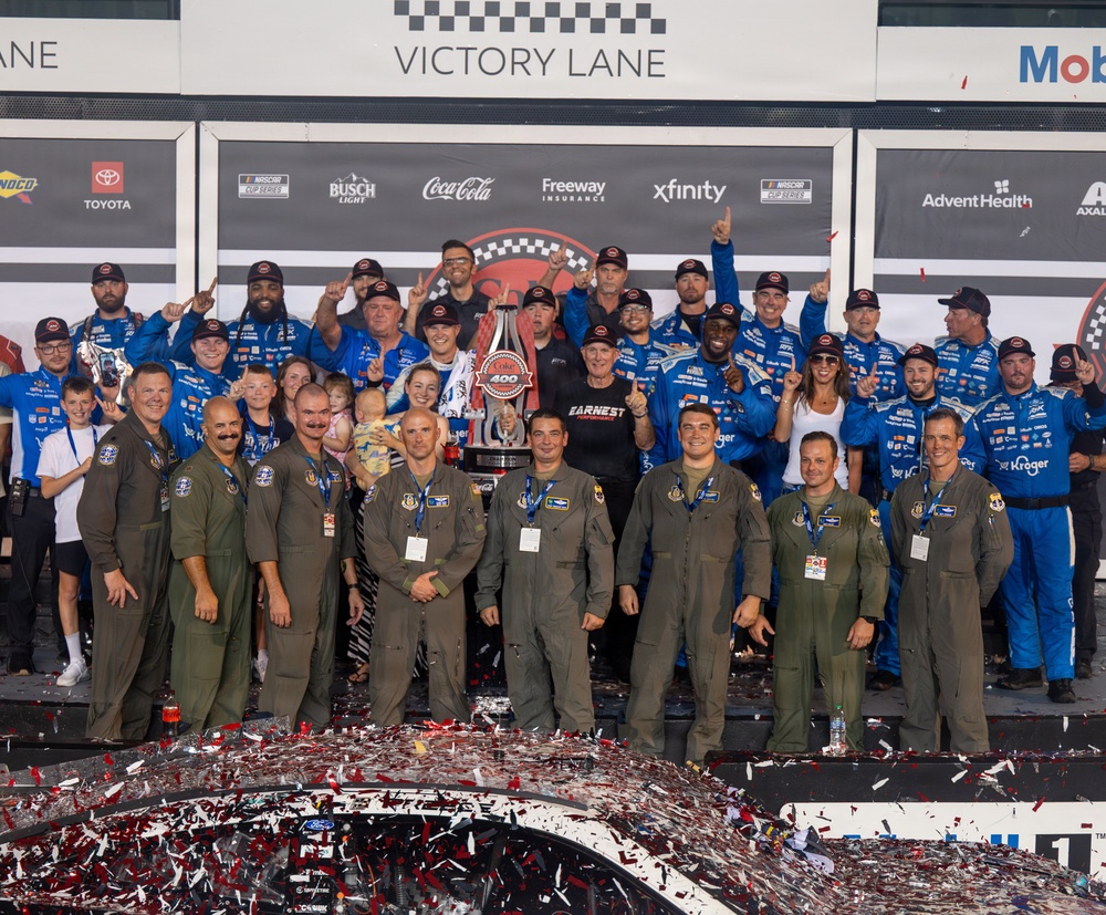 A large group celebrates a NASCAR Cup Series victory in Victory Lane, with racing team members in blue uniforms and military personnel in olive flight suits posing together. A trophy is visible at the center of the group, and confetti covers the foreground along with the roof of a race car. Sponsor banners including Toyota, Busch Light, Coca-Cola, Freeway Insurance, Xfinity, and AdventHealth are visible in the background.