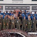 A large group celebrates a NASCAR Cup Series victory in Victory Lane, with racing team members in blue uniforms and military personnel in olive flight suits posing together. A trophy is visible at the center of the group, and confetti covers the foreground along with the roof of a race car. Sponsor banners including Toyota, Busch Light, Coca-Cola, Freeway Insurance, Xfinity, and AdventHealth are visible in the background.
