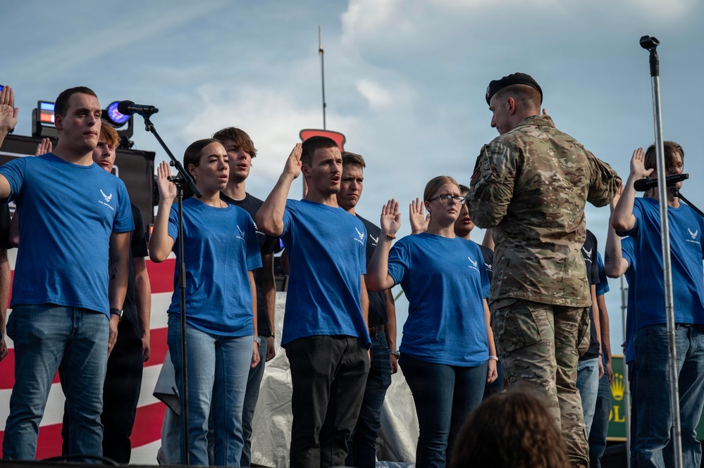 A military enlistment or oath-of-office ceremony is taking place on a public stage. A uniformed soldier in MultiCam/OCP camouflage and beret faces a group of recruits wearing blue U.S. Air Force t-shirts, all with their right hands raised. The ceremony appears to be conducted at a public event, evidenced by the stage setup, microphones, and a Rolex advertisement banner visible in the background.