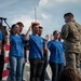 A military enlistment or oath-of-office ceremony is taking place on a public stage. A uniformed soldier in MultiCam/OCP camouflage and beret faces a group of recruits wearing blue U.S. Air Force t-shirts, all with their right hands raised. The ceremony appears to be conducted at a public event, evidenced by the stage setup, microphones, and a Rolex advertisement banner visible in the background.
